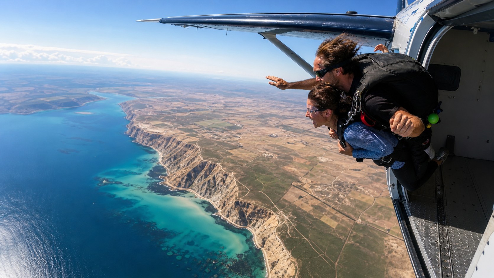 Tandem skydivers exiting an aircraft above a bright turquoise coastline